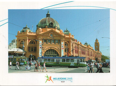 A Melbourne tram passes in front of the iconic Flinders Street Station building, a historic architectural landmark with a prominent dome, under a clear blue sky, with people on the street.