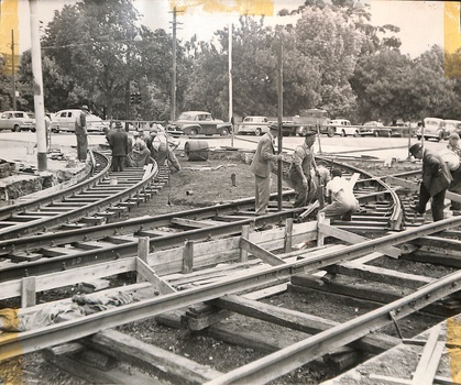Men in work clothes and hats construct tram tracks on a dirt construction site with vintage cars in the background.
