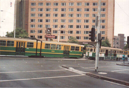 Two yellow and green trams at a street intersection in front of a tall building with many windows and a Travelodge sign.