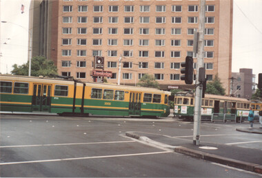 Two yellow and green trams at a street intersection in front of a tall building with many windows and a Travelodge sign.