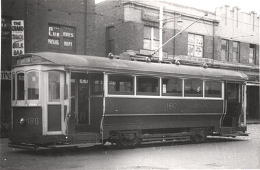 A vintage tram, number 180, sits parked on a street in front of brick buildings with storefront signs.