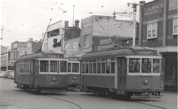 Three vintage trams are stopped on a city street, with passengers visible inside. Buildings with various advertisements line the street behind the trams.