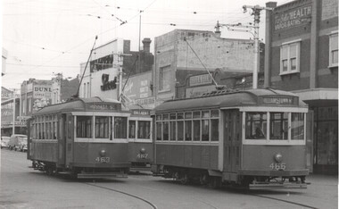 Three vintage trams are stopped on a city street, with passengers visible inside. Buildings with various advertisements line the street behind the trams.
