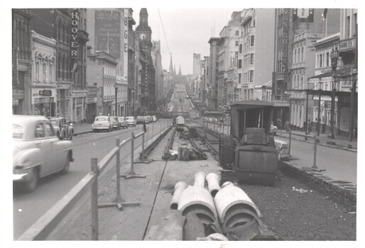 A street scene with tram tracks being laid in the foreground. Cars drive on the left side of the street, and buildings line both sides. Construction equipment and materials are visible near the tracks.