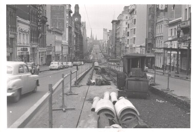 A street scene with tram tracks being laid in the foreground. Cars drive on the left side of the street, and buildings line both sides. Construction equipment and materials are visible near the tracks.