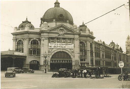 Shows the grand facade of a large, ornate building with multiple domes and a central clock tower. In front of the building, several vintage cars are parked on a street, and a group of men appear to be working on road construction. A large banner across the building reads "DON'T USE THE WORD 'MAGNIFICENT' UNTIL YOU'VE SEEN MT. BUFFALO NATIONAL PARK."