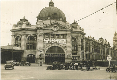 Shows the grand facade of a large, ornate building with multiple domes and a central clock tower. In front of the building, several vintage cars are parked on a street, and a group of men appear to be working on road construction. A large banner across the building reads "DON'T USE THE WORD 'MAGNIFICENT' UNTIL YOU'VE SEEN MT. BUFFALO NATIONAL PARK."