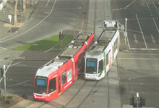 Two modern trams, one red and one white with green accents, sit side-by-side at a tram junction on a city street.