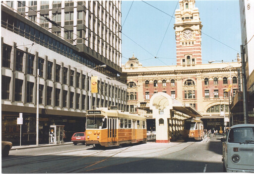 A yellow tram is arriving at a terminus in front of a large, modern building and a historic building with a clock tower. The tram has the number 153 on its front.