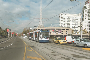 A modern, blue and white tram travels down a city street lined with buildings, with a distinctive spire in the background.