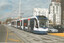 A modern, blue and white tram with "SIEMENS" branding and advertising for "PLUS" is arriving at a tram stop on a city street. The tram is part of a multi-car train, with additional white trams visible further down the tracks. In the background, tall buildings and a distinctive white spire-like structure are visible under a cloudy sky. 