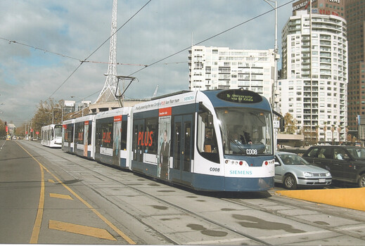 A modern, blue and white tram with "SIEMENS" branding and advertising for "PLUS" is arriving at a tram stop on a city street. The tram is part of a multi-car train, with additional white trams visible further down the tracks. In the background, tall buildings and a distinctive white spire-like structure are visible under a cloudy sky. 
