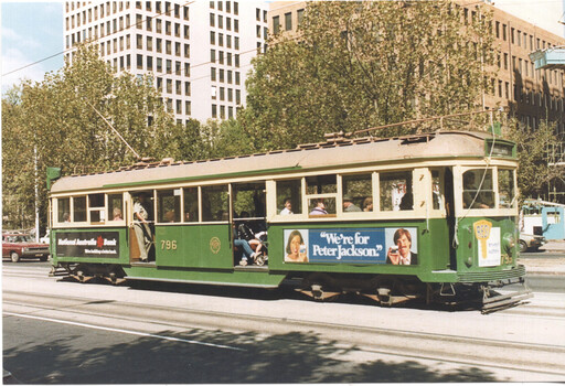 A vintage green and cream tram numbered 796 travels along a city street, with passengers visible inside through the windows. An advertisement for "Peter Jackson" cigarettes is prominently displayed on the side of the tram, alongside advertising for National Australia Bank. Tall buildings and leafy trees line the street in the background, suggesting an urban environment.