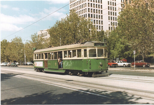 A vintage green and cream tram numbered 772 travels along tram tracks on a city street, with trees and a tall modern building in the background.