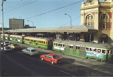 Several green and yellow trams are lined up on a track in front of a large, ornate building with a dome, alongside a road with cars.