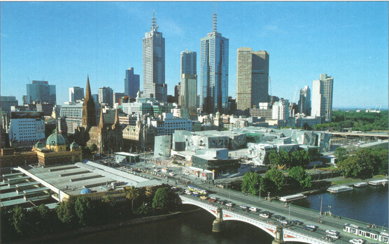 Panoramic view of a cityscape featuring a river in the foreground with a bridge, and a dense collection of modern skyscrapers and historical buildings in the background under a clear blue sky.