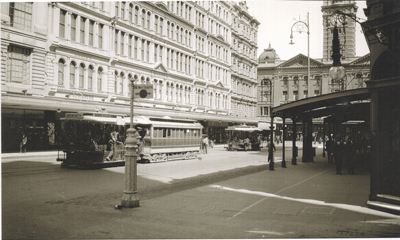 Street scene with multiple cable cars and pedestrians in front of ornate, multi-story buildings.