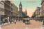 A busy street scene in Melbourne, Australia, with ornate 19th-century buildings lining the sides. A prominent clock tower rises above the architecture. Several cable trams are visible on the street, carrying passengers. Pedestrians stroll along the footpaths.