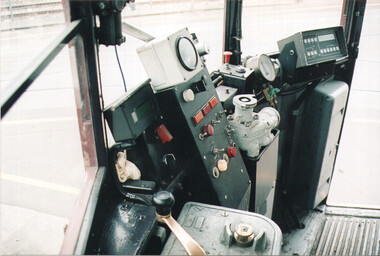 Close-up view of a complex and cluttered tram driver's cabin, showing various gauges, levers, buttons, and a digital display on a dark panel.