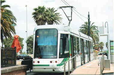 A modern white tram numbered 3028 with green accents is stopped at a tram stop. The tram's destination display reads "109 BOX HILL CITY". Palm trees and a cloudy sky are visible in the background.