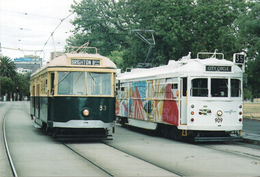 Two historic trams on tram tracks; one is dark green and cream with "BRIGHTON BCH" on the front and number 53, the other is white with colorful abstract art and "CITY CIRCLE" on the front with number 909.