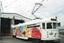 A white, vintage electric tram decorated with a colorful, abstract mural on its side, stands in front of an open-doored industrial building.