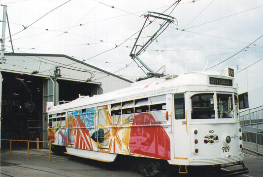 A white, vintage electric tram decorated with a colorful, abstract mural on its side, stands in front of an open-doored industrial building.