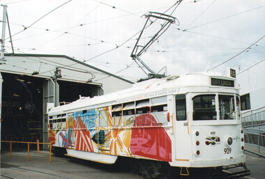 A white, vintage electric tram decorated with a colorful, abstract mural on its side, stands in front of an open-doored industrial building.