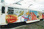 A white tram is decorated with a colorful mural celebrating "100 years of electric trams" and "Melbourne all over". The artwork features stylized representations of older trams, books, birds, and abstract shapes in bold reds, blues, yellows, and oranges. Text on the side of the tram reads "Yarra Trams" and "Meet you in the heart of the city".