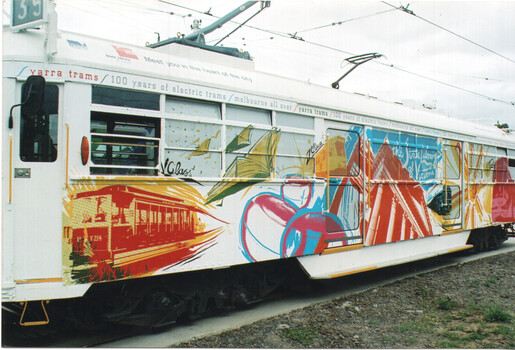 A white tram is decorated with a colorful mural celebrating "100 years of electric trams" and "Melbourne all over". The artwork features stylized representations of older trams, books, birds, and abstract shapes in bold reds, blues, yellows, and oranges. Text on the side of the tram reads "Yarra Trams" and "Meet you in the heart of the city".