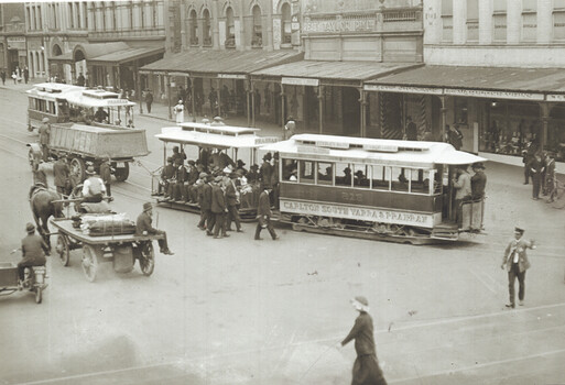 A busy city street scene with horse-drawn carts and cable trams, showing passengers riding on open-sided tram cars labeled "Carlton South Yarra & Prahran."  One of the cable trams is being towed by a truck.