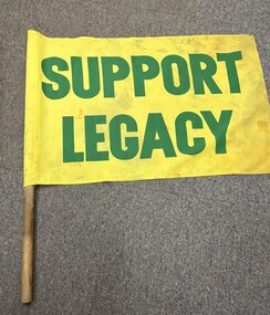 A yellow flag with green lettering reading "SUPPORT LEGACY" held by a wooden pole.