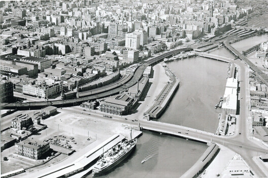 Aerial view of the Yarra River with a large passenger ship docked alongside piers and warehouses, surrounded by a dense urban cityscape with railway lines and three bridges.