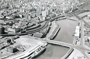 Aerial view of the Yarra River with a large passenger ship docked alongside piers and warehouses, surrounded by a dense urban cityscape with railway lines and three bridges.