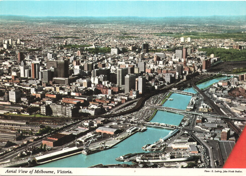 Aerial view of a dense city skyline with numerous tall buildings, a winding river or waterway with bridges, and a harbor area with ships and cranes.