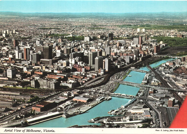 Aerial view of a dense city skyline with numerous tall buildings, a winding river or waterway with bridges, and a harbor area with ships and cranes.