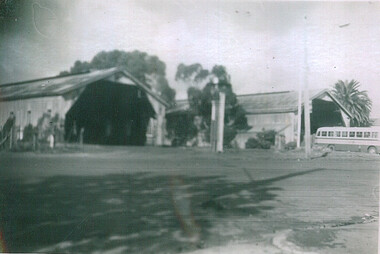 Shows several long, low buildings with gabled roofs and open entrances. A white and light-colored bus is parked to the right of the buildings. Palm trees and other foliage are visible. 