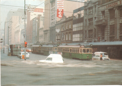Flooded city street with trams and submerged vehicles with traffic lights and buildings.