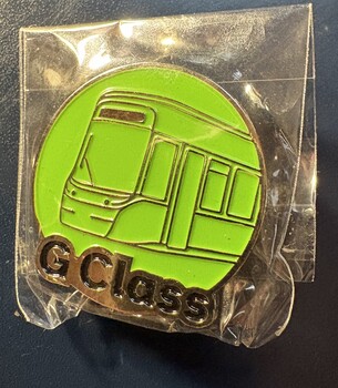 A circular, lime-green enamel pin with a gold outline depicting the front of a modern tram, featuring large windows. The words "G Class" are embossed in gold text below the tram. The pin is enclosed in clear plastic packaging.