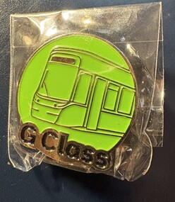 A circular, lime-green enamel pin with a gold outline depicting the front of a modern tram, featuring large windows. The words "G Class" are embossed in gold text below the tram. The pin is enclosed in clear plastic packaging.