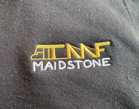 Embroidered logo on dark fabric featuring stylized yellow and white text "TMF MAIDSTONE." The logo combines bold yellow letters with a white vertical text element, indicating branding or affiliation with Maidstone location.