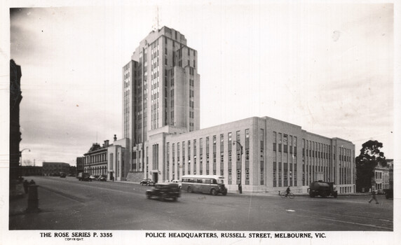 A panoramic view of a large, imposing police headquarters building with a tall tower and a long, horizontal section, located on a street with vehicles and pedestrians.