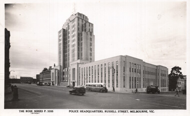 A panoramic view of a large, imposing police headquarters building with a tall tower and a long, horizontal section, located on a street with vehicles and pedestrians.