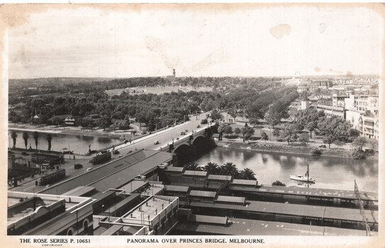 Panoramic view from above, showing Princes Bridge in Melbourne spanning a wide river, with tram cars and vehicles on the bridge, and a boat on the water. Buildings and lush trees line the riverbanks, with a railway station visible at the bottom of the card.