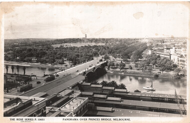 Panoramic view from above, showing Princes Bridge in Melbourne spanning a wide river, with tram cars and vehicles on the bridge, and a boat on the water. Buildings and lush trees line the riverbanks, with a railway station visible at the bottom of the card.
