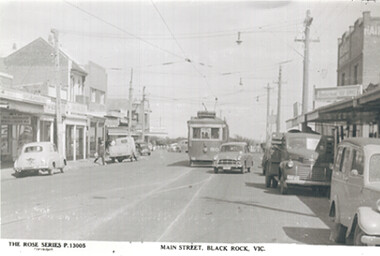 A vintage, black and white photograph depicting a bustling main street in Black Rock, Victoria, featuring a tram, various cars, and shops along the footpaths under a bright, hazy sky.