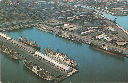 Aerial view of a busy harbor - Melbourne - filled with cargo ships docked along multiple piers and industrial buildings.