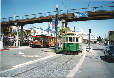 Two vintage trams, one red and one green, on tracks with a pedestrian bridge overhead labeled "PIER 39" in San Francisco.  