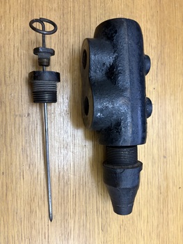 Black cast metal valve body with two ports on the left and a threaded nozzle on the right, shown above a disassembled plunger with a spring clip, threaded housing, and a long metal rod.