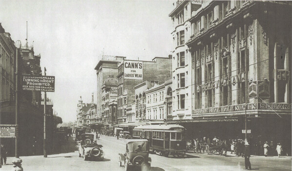 A busy city street with vintage cars and trams, flanked by multi-story buildings with shops and signs including "Cann's for Ladies Wear" and "Square Deal Sale Now On".